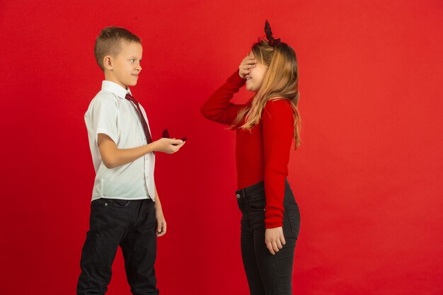 Genau wie ein Erwachsener. Valentinstagfeier, glückliche, niedliche kaukasische Kinder lokalisiert auf rotem Studiohintergrund. Konzept der menschlichen Gefühle, Gesichtsausdruck, Liebe, Beziehungen, romantische Feiertage.