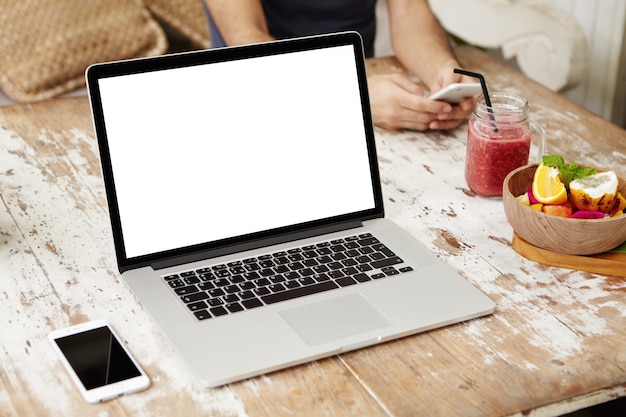 Generischer moderner Laptop mit leerem Kopierraumbildschirm, der auf Holztisch mit Handy, Smoothie und Früchten ruht.