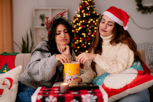 genervte hübsche junge Mädchen mit Weihnachtsmütze und Stechpalmenkranz essen und schauen sich den Popcorn-Eimer an, der zu Weihnachten auf Sesseln zu Hause sitzt