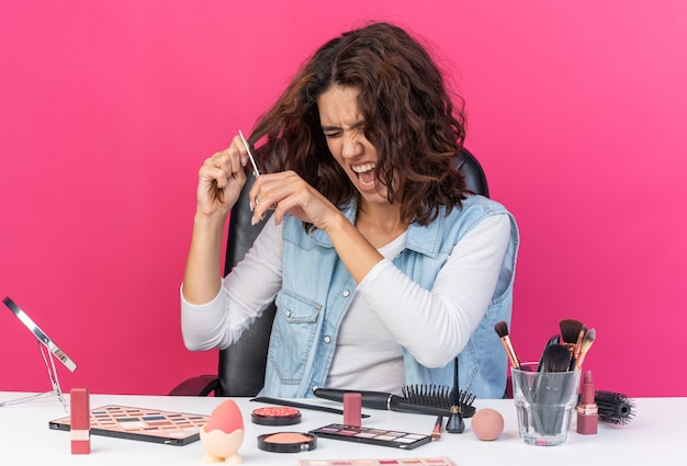 Genervte hübsche kaukasische Frau, die am Tisch mit Make-up-Werkzeugen sitzt und ihr Haar mit einer Schere schneidet, isoliert auf rosa Wand mit Kopierraum