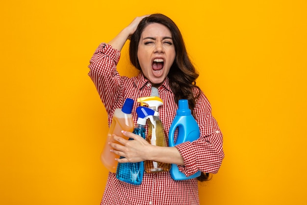 Genervte hübsche kaukasische Putzfrau, die Reinigungssprays und -flüssigkeiten hält und sich die Hand auf den Kopf legt