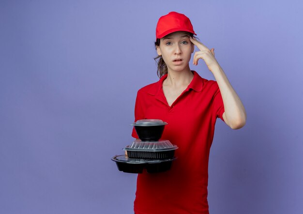 Genervte junge hübsche Lieferfrau, die rote Uniform und Kappe hält, die Lebensmittelbehälter hält, die Selbstmordgeste tun, lokalisiert auf lila Hintergrund mit Kopienraum