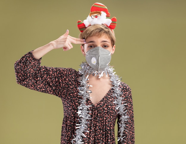 Kostenloses Foto genervtes junges hübsches mädchen, das weihnachtsmann-stirnband und lametta-girlande um den hals mit schutzmaske trägt, die kamera betrachtet, die selbstmordgeste tut, lokalisiert auf olivgrünem hintergrund