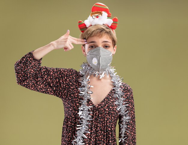 Genervtes junges hübsches Mädchen, das Weihnachtsmann-Stirnband und Lametta-Girlande um den Hals mit Schutzmaske trägt, die Kamera betrachtet, die Selbstmordgeste tut, lokalisiert auf olivgrünem Hintergrund