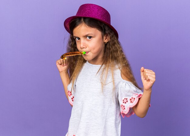 genervtes kleines kaukasisches Mädchen mit violettem Partyhut, der Partypfeife bläst und Fäuste isoliert auf lila Wand mit Kopienraum hält