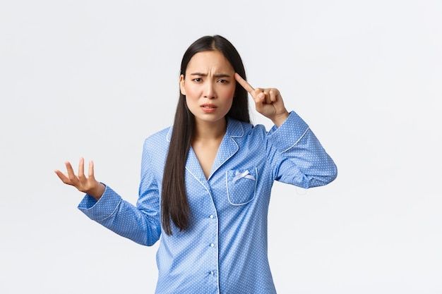 Genervtes und verwirrtes asiatisches Mädchen, das sich darüber beschwert, dass sich jemand dumm oder verrückt verhält, blaue Pyjamas trägt, frustriert die Hand hebt und mit dem Finger vor Entsetzen über die Schläfe rollt, jemanden schimpft