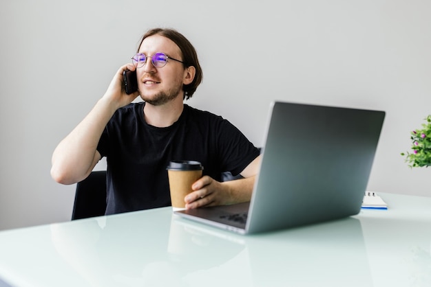 Genießen Sie einen guten Arbeitstag Selbstbewusster junger Mann, der am Laptop arbeitet und am Handy spricht, während er an seinem Arbeitsplatz im Büro sitzt