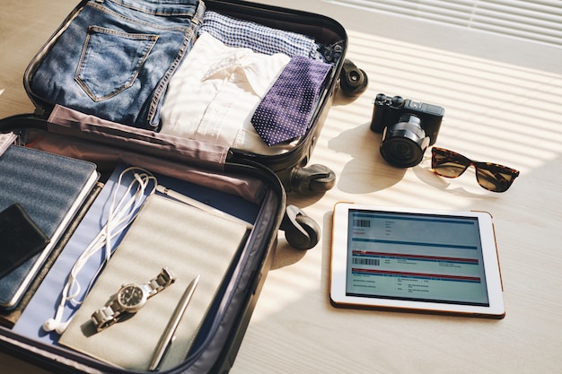 Gepackter Koffer auf dem Schreibtisch, Tablet mit Eticket auf dem Bildschirm, Kamera und Sonnenbrille