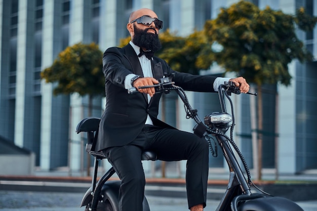 Gepflegter moderner Geschäftsmann mit Sonnenbrille fährt Elektroroller zu seinem Büro.