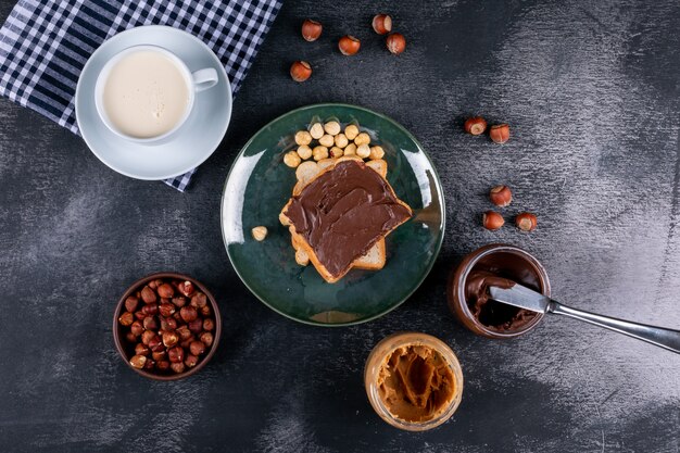 Gereinigte und geschälte Haselnüsse in einem grünen Glasteller mit Kakaobrot, Milch flach auf einem dunklen Steintisch