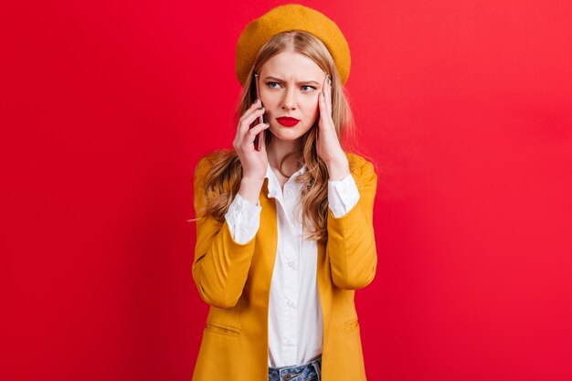 Gereizte blonde Frau in der gelben Baskenmütze, die am Telefon spricht. kaukasisches Mädchen in der Jacke, die Smartphone an der roten Wand hält.