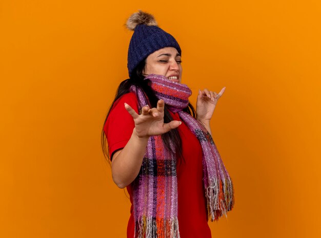 Gereizte junge kranke Frau, die Wintermütze und Schal trägt, die in der Profilansicht stehen und Hände in Luft mit geschlossenen Augen halten, die auf orange Wand lokalisiert werden