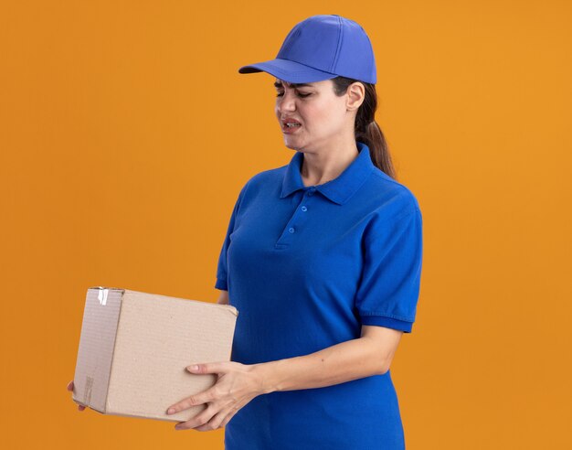 Gereizte junge Lieferfrau in Uniform und Mütze, die in der Profilansicht steht und Karton isoliert auf oranger Wand mit Kopierraum hält und betrachtet