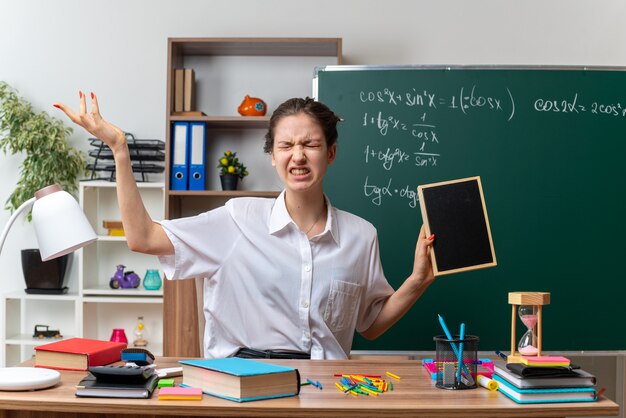 Gereizte junge Mathematiklehrerin, die am Schreibtisch mit Schulmaterial sitzt und eine Mini-Tafel hält, die leere Hand mit geschlossenen Augen im Klassenzimmer zeigt