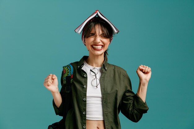 Gereizte junge Studentin, die Kopftuch und Rucksack trägt und eine Brille auf ihre Bluse setzt, die Fäuste in der Luft hält und einen offenen Notizblock auf dem Kopf hält, mit geschlossenen Augen isoliert auf blauem Hintergrund