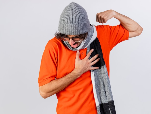 Kostenloses Foto gereizter junger kaukasischer kranker mann, der brille wintermütze und schal tut, die starke geste tut, die hand auf brust hält, die zunge lokalisiert auf weißem hintergrund mit kopienraum zeigt