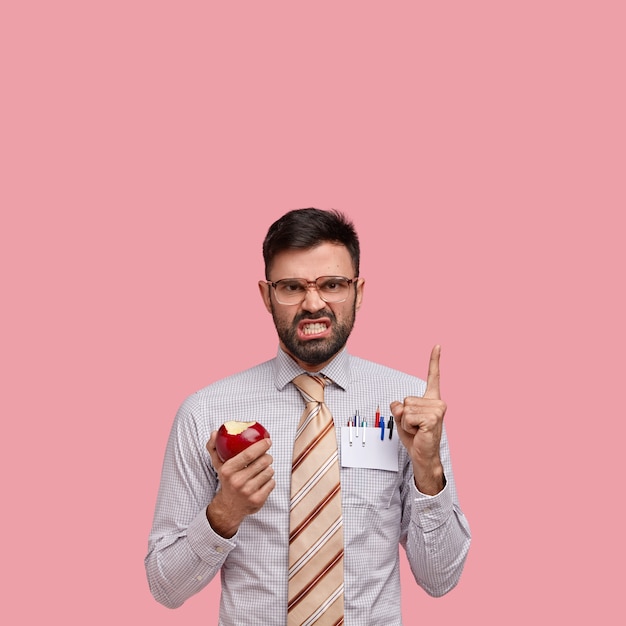 Kostenloses Foto gereizter unrasierter unternehmer zeigt auf eine lücke oben, runzelt unzufrieden die stirn, trägt eine große brille und isst roten apfel