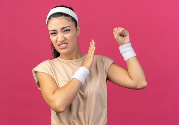 Gereiztes junges kaukasisches sportliches Mädchen mit Stirnband und Armbändern, das die Faust in der Luft hält und die Verweigerungsgeste isoliert auf rosa Wand macht