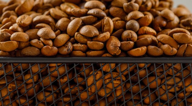 Geröstete Kaffeebohnen in einem schwarzen Korb auf einer leicht weißen Oberfläche. Seitenansicht.