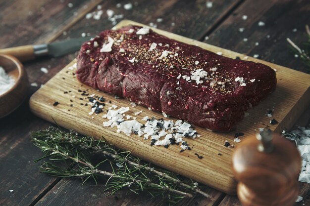 Gesalzenes, gepfeffertes Stück Fleisch mit Fokus, bereit zum Grillen auf einem Holztisch zwischen Kräutern