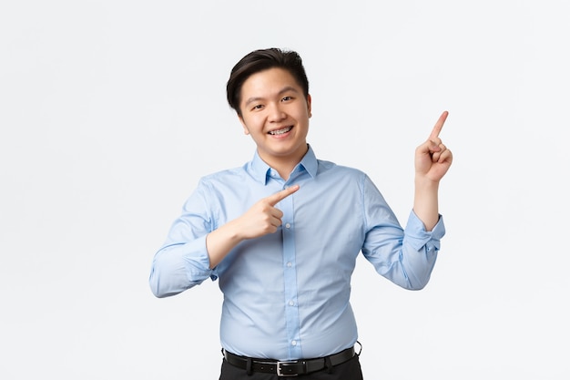Geschäfts-, Finanz- und Menschenkonzept. Angenehm lächelnder asiatischer Verkäufer in blauem Hemd, Zahnspangen, Finger zeigen in der oberen rechten Ecke, Ankündigung machen, Diagramm oder Produkt anzeigen, weißer Hintergrund.