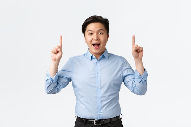 Geschäfts-, Finanz- und Menschenkonzept. Aufgeregter und erstaunter asiatischer Geschäftsmann in blauem Hemd, der Ankündigung macht, mit den Fingern nach oben zeigt und in die Kamera schaut, große Neuigkeiten erzählt, weißer Hintergrund.