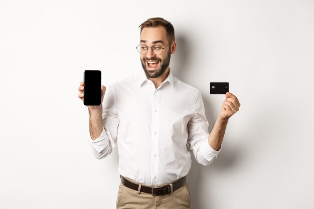 Geschäfts- und Online-Zahlung. Lächelnder hübscher Mann, der mobilen Bildschirm und Kreditkarte zeigt, stehend