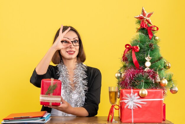 Geschäftsdame im Anzug mit Brille, die ihr Geschenk zeigt, das Brillengesten macht und an einem Tisch mit einem Weihnachtsbaum darauf im Büro sitzt