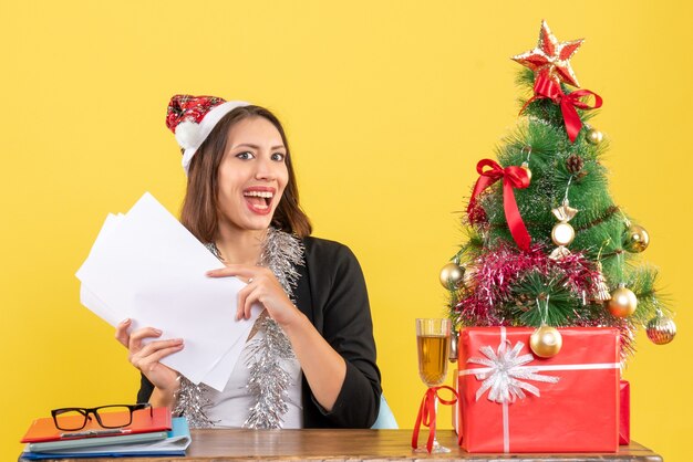 Geschäftsdame im Anzug mit Weihnachtsmannhut und Neujahrsdekorationen, die allein arbeiten und an einem Tisch mit einem Weihnachtsbaum darauf im Büro sitzen