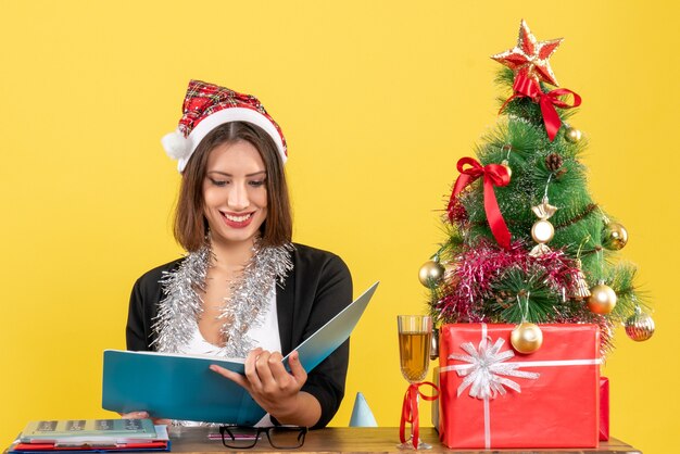 Geschäftsdame im Anzug mit Weihnachtsmannhut und Neujahrsdekorationen, die Dokument lesen und an einem Tisch mit einem Weihnachtsbaum darauf im Büro sitzen