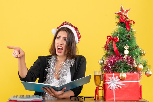 Geschäftsdame im Anzug mit Weihnachtsmannhut und Neujahrsdekorationen, die Dokument überprüfen, das etwas zeigt und an einem Tisch mit einem Weihnachtsbaum darauf im Büro sitzt