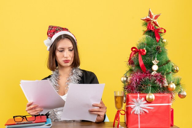 Geschäftsdame im Anzug mit Weihnachtsmannhut und Neujahrsdekorationen, die Dokumente überprüfen und an einem Tisch mit einem Weihnachtsbaum darauf im Büro sitzen