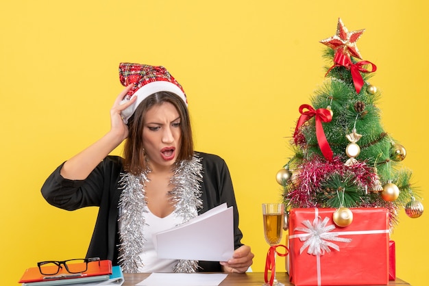 Geschäftsdame im Anzug mit Weihnachtsmannhut und Neujahrsdekorationen, die sich nervös fühlen und an einem Tisch mit einem Weihnachtsbaum darauf im Büro sitzen