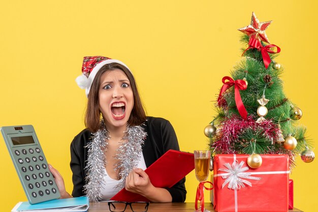 Geschäftsdame im Anzug mit Weihnachtsmannhut und Neujahrsdekorationen, die sich verwirrt fühlen, während sie Buchhaltung überprüfen und an einem Tisch mit einem Weihnachtsbaum darauf im Büro sitzen