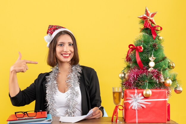 Geschäftsdame im Anzug mit Weihnachtsmannhut und Neujahrsdekorationen, die sich zeigen und an einem Tisch mit einem Weihnachtsbaum darauf im Büro sitzen