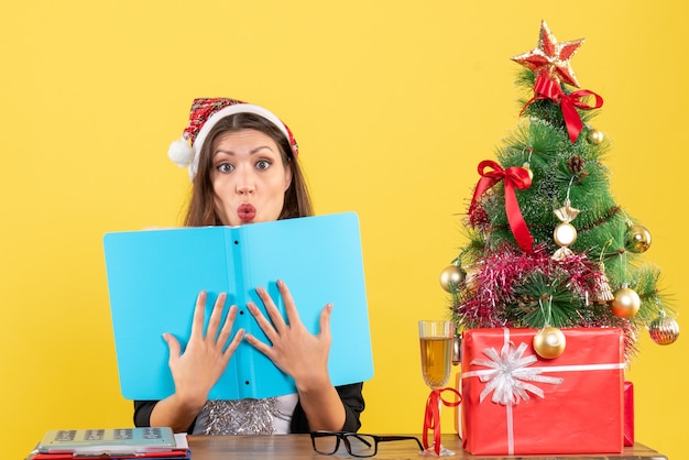 Geschäftsdame im Anzug mit Weihnachtsmannhut und Neujahrsdekorationen konzentrierte sich auf Dokument in tiefen Gedanken und saß an einem Tisch mit einem Weihnachtsbaum darauf im Büro