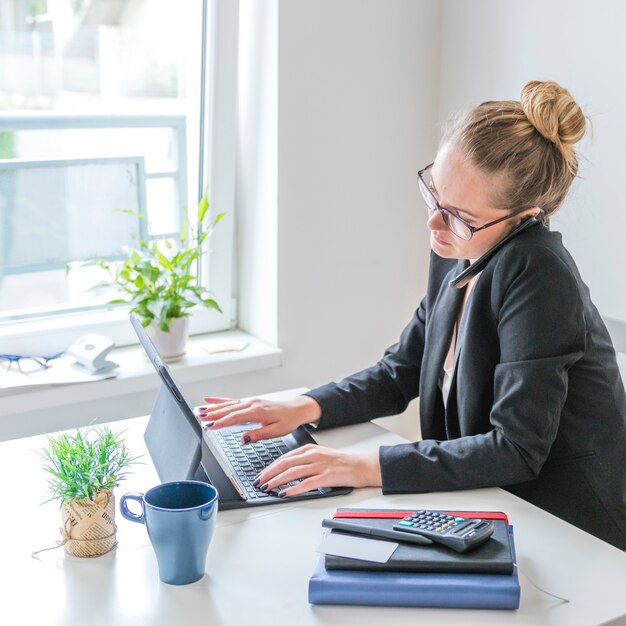 Geschäftsfrau, die an Laptop unter Verwendung des Mobiltelefons im Büro arbeitet