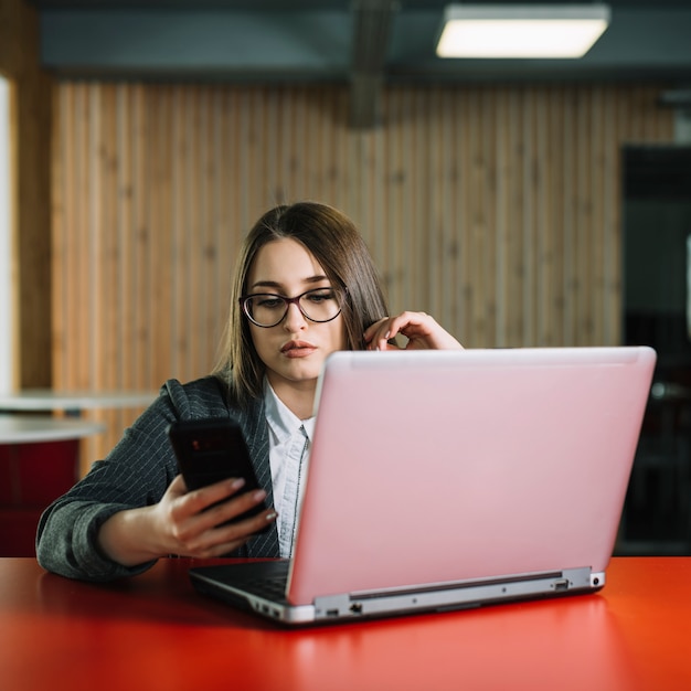 Geschäftsfrau, die bei Tisch Smartphone mit Laptop verwendet