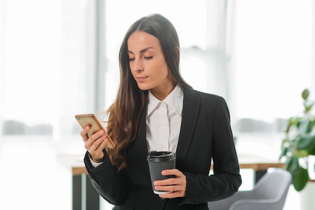 Geschäftsfrau, die den Handy hält Wegwerfkaffeetasse betrachtet