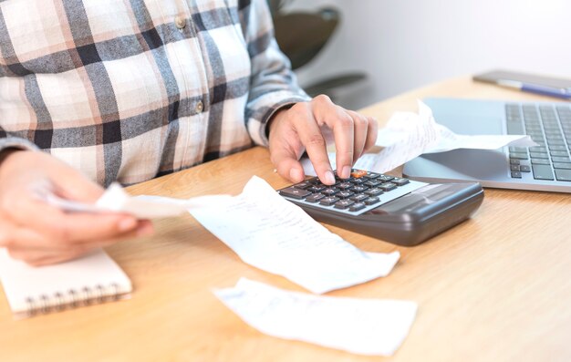 Geschäftsfrau, die den Rechner drückt, berechnet die verschiedenen Kosten, die durch die gehaltenen und auf den Tisch gelegten Rechnungsrechnungen bezahlt werden müssen.