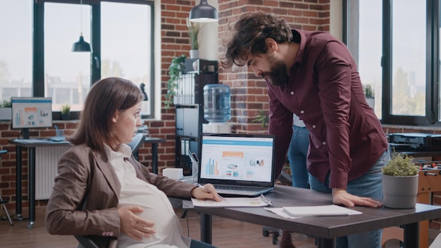Geschäftsfrau, die ein Kind erwartet und an der Analyse mit einem Kollegen für die Projektplanung arbeitet. Mann, der schwangeren Mitarbeitern bei der Wachstums-, Entwicklungs- und Marketingstrategie auf dem Laptop hilft.