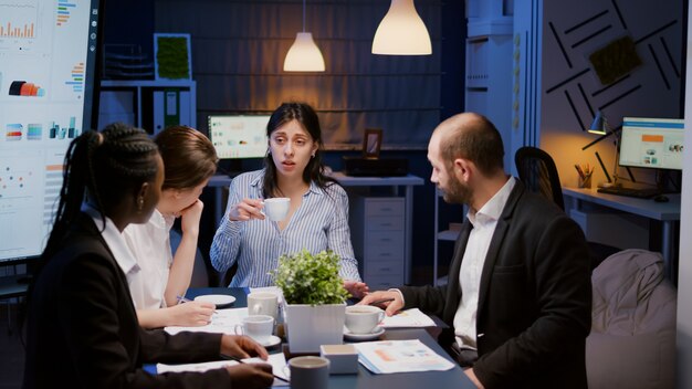 Geschäftsfrau, die eine Tasse Kaffee hält, während sie mit multiethnischer Teamarbeit diskutiert, die ein Managementprojekt mit Diagrammpapieren löst. Diverse Kollegen, die spät in der Nacht im Besprechungsraum arbeiten