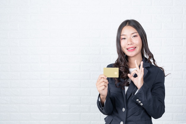 Geschäftsfrau, die eine unterschiedliche Geldautomatenkarte, weiße Backsteinmauer hält Machte Gesten mit Gebärdensprache.