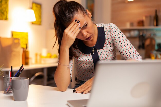 Geschäftsfrau, die erschöpft aussieht und die Stirn massiert, während sie Überstunden auf dem Laptop aus der heimischen Küche macht. Mitarbeiter, die um Mitternacht moderne Technologie verwenden, machen Überstunden für Job, Geschäft, Karriere, Netzwerk