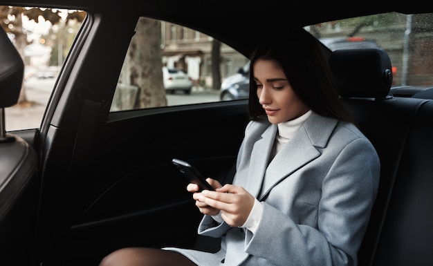 Geschäftsfrau, die mit dem Auto auf dem Rücksitz reist und Textnachricht auf Smartphone liest, während sie auf Besprechung fährt