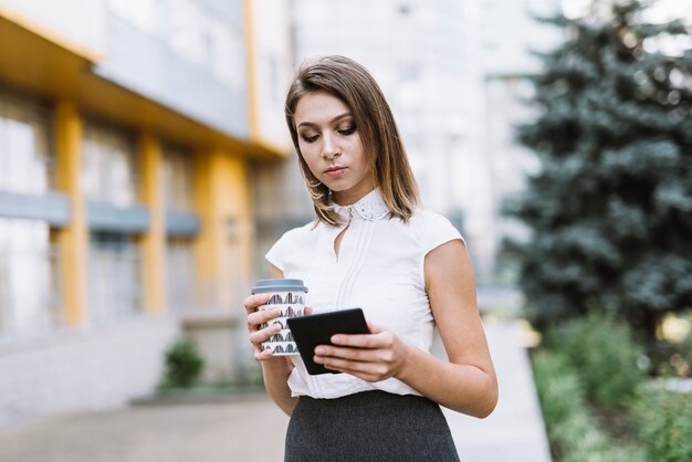 Geschäftsfrau, die Mitnehmerkaffeetasse betrachtet Smartphone betrachtet