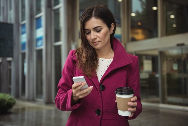 Geschäftsfrau, die wegwerfbare Kaffeetasse hält und Handy verwendet