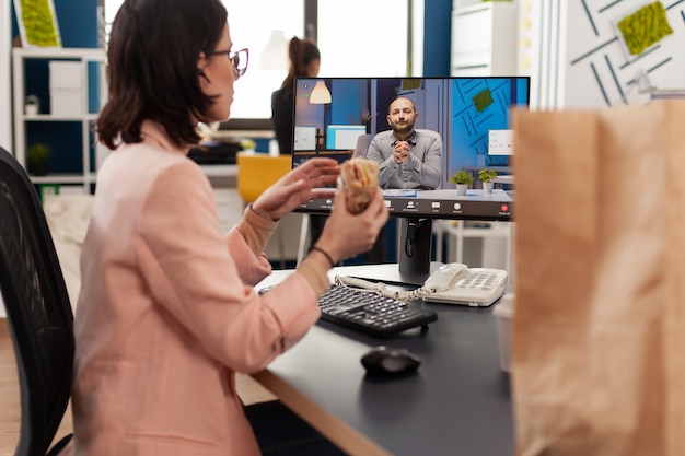 Geschäftsfrau isst Liefer-Sandwich zum Mitnehmen während einer Online-Videokonferenz-Konferenz, die mit einem Remote-Mitarbeiter diskutiert?