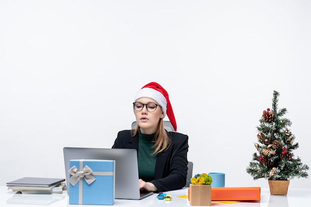 Geschäftsfrau mit einem Weihnachtsmannhut, der an einem Tisch mit einem Weihnachtsbaum und einem Geschenk sitzt