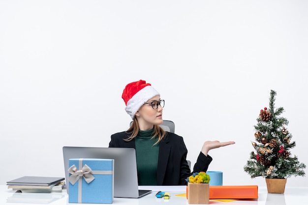 Geschäftsfrau mit einem Weihnachtsmannhut, der an einem Tisch mit einem Weihnachtsbaum und einem Geschenk sitzt